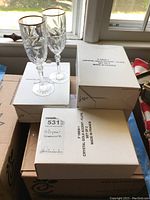 Eight crystal flute glasses with gold rims stacked in boxes labeled 'Crystal Gold Accent - Flute Made in France'. Two glasses are shown standing on boxes.