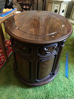 Round wooden end table with a visible wood grain top and circular inlay, showing surface chips and scratches. Table has paneled doors beneath the top, with decorative handles and a deep wood finish.
