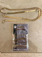 Flat 14k gold herringbone/chevron patterned chain laid out next to packaged Nicole gold plated Figaro chain