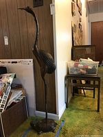 Full view of tall metal sculpture of crane standing on turtle base against wood panel wall.