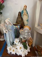 Front view showing five religious figurines on wooden base with white artificial flowers, including two large Virgin Mary figures, one dark statue, glass lamp and small wooden statue.