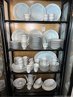 Shelf displaying multiple pieces of white china with floral and gold detailing including plates, cups, saucers, creamers, pitchers, serving bowls, and a gravy boat.