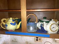 Shelf displaying five tea pots and a tea pot clock, arranged side by side with visible wear on the glass pot handle.