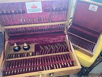 Two open wooden boxes with plush burgundy velvet lining, each containing a complete set of bronze Thai fondue forks and flatware including forks, knives, spoons, and a pair of small condiment shakers in one box.