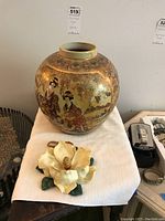 Front view of a large Asian ceramic vase featuring two traditional figures in elaborate costumes, resting on a white cloth, with a cream-colored ceramic flower sculpture beside it.