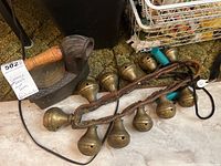 Photo showing ten vintage brass sleigh bells attached to two braided leather straps and a vintage cast iron piece with wooden handle on carpeted floor.