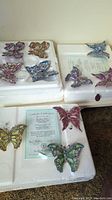Top and angled view of multiple porcelain butterfly ornaments in white foam trays, showing variety of colors and certificates of authenticity beneath some butterflies.