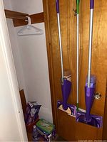 Full view of two Swiffer Wet Jet mops leaning against wooden door with boxes of refills and bucket on floor