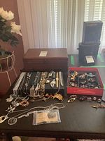 Full view showing wooden jewelry box and multiple jewelry pieces spread out on the table including chains, brooches, watches, cufflinks, tie tack, and faux pearls.