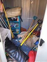 View inside storage shed showing snow shovels with plastic blades, red gas cans, purchase bags, and other stored outdoor tools.