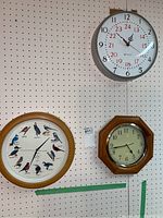 Three wall clocks hanging on pegboard wall. One is an Edwards 24-hour clock with white face, one is a wood framed bird illustrated clock, and the third is a wood framed hexagonal clock with white face.