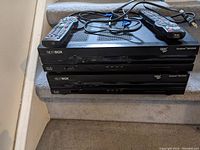 Photo of two Rogers NextBox Explorer 8642HD PVR devices stacked on carpeted stairs with two remote controls and cables.