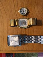 Photo of three watches on a wooden surface: one ladies watch head without strap, two men's watches with metal straps (gold-tone expandable and silver-tone link bracelet)