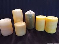 Six pillar candles in yellow, off white, and apple green colors, against a dark blue background.