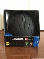 Photo of black Bollé snow helmet in original box on hardwood floor. Size marked L-G (58-60cm). Shows front and top vents.