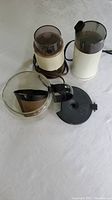 Two Braun electric coffee grinders, a clear glass coffee pot with black handle, and a black plastic filter holder with lid laid out on a white surface.
