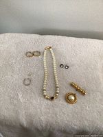 Costume jewelry items including pearl-like necklace with blue centerpiece, gold hoops, black studs, and brooches arranged on fabric