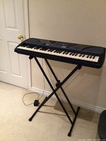 Full view of Yamaha electronic keyboard mounted on black X-style stand with power adapter cable visible.
