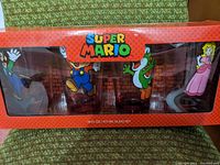 Boxed set showing all 4 Super Mario character glasses visible through transparent window. Box is red with Super Mario logo on top.