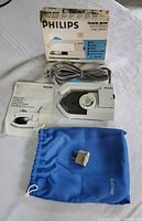 Philips travel iron displayed with original box, instruction manual, plug adapter, and blue carry bag on a white cloth background.