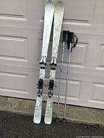 Full view of pair of white and grey patterned women's skis standing next to silver-blue ski poles with black handles.