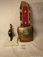 Cowbell with decorative strap and lidded pipe placed on cloth background