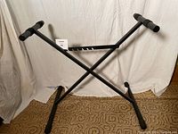 Black metal X-frame adjustable electronic piano stand shown unfolded with keyboard support arms and adjustable height notches visible.