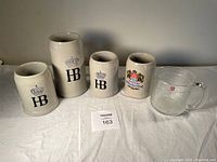 Four stoneware Hofbrauhaus beer steins and one clear Iittala glass mug on white surface, showing front logos and sizes.