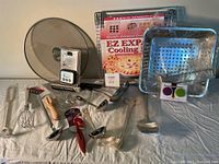 Various kitchen tools including ladles, whisk, barbecue basket, cooling rack, glass pot lid, and digital timer arranged on white cloth