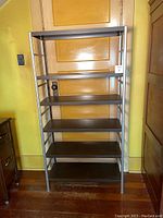 Front view of the tall metal shelving unit with five faux wood shelves against a yellow wall and closed door.