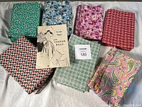 Seven assorted cotton feed sack fabrics in various floral, checkered, and paisley prints along with the vintage 'Needle Magic with Cotton Bags' booklet.