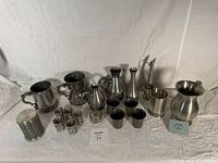 Group view of multiple pewter items including containers, cups, pitchers, and vases laid out against a white backdrop.