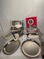Set of stainless steel serving dishes displayed on white fabric, includes round plates and oval serving dishes, some boxed.