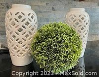 Pair of white ceramic candle holders with geometric lattice cut-out design and a round faux green foliage decor ball in between, displayed against stone background.