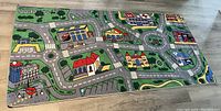 Full view of the rectangular children's carpet featuring a painted town scene with roads, parking, airport, bus stop, buildings, and greenery on a hardwood floor.