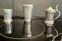 Three pewter miniature goblets arranged side by side showing different embossed designs and one with hinged lid and handle.