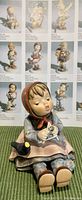Photo of the Hummel figurine "Happy Pastime" showing a seated child holding a bird and paintbrush in detail, placed on a textured green surface with a catalog background.