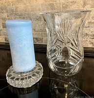 Two clear lead crystal hurricane candle holders shown on a black surface with a beige tiled background. One holder contains a blue candle. Both pieces are heavy and intricately cut.
