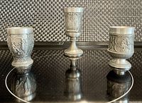 Photo of three miniature pewter goblets with embossed decorative motifs standing on a reflective black surface.