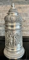 Front side of pewter stein showing detailed relief scene with groups of people and text on base