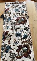 Full view of the floral-patterned table runner on a wooden table, showing cream base color with teal, brown, and mustard floral designs.