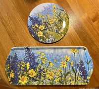 Round teapot stand and rectangular sandwich tray with matching floral designs in yellow, purple, and blue tones, on wooden surface.