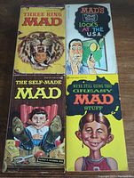 Photo of four Mad magazine pocketbooks arranged on wooden surface, covers showing Alfred E. Neuman and other classic cover illustrations.