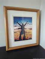 Photo of the entire framed artwork showing the silhouette figure with rings against a sunset background, in light wood frame.