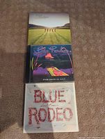 Three Blue Rodeo CD cases stacked showing their front covers: The Things We Left Behind at top, Five Days in July middle, Small Miracles bottom.