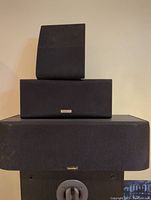 Three speakers stacked vertically against a wall, Paradigm center channel speaker on bottom, Kenwood speaker in middle, and smaller speaker on top.