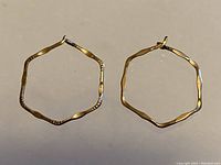 Pair of hexagonal shaped 10K gold hoop earrings shown flat on a neutral surface, clearly showing their shape, color, and texture.
