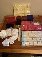 Photo showing collection of various jewelry boxes in cream, red, blue colors with bows, several hinged lidded cases and dozen hand-crocheted white cotton drawstring bags arranged on wooden table.