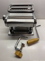 Front view of stainless steel Grazia pasta maker with two detachable hand cranks and clamp on white surface.