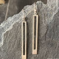 Front view photo of the pair of elongated rectangular sterling silver earrings with a hollow center cutout, placed on a dark textured surface.